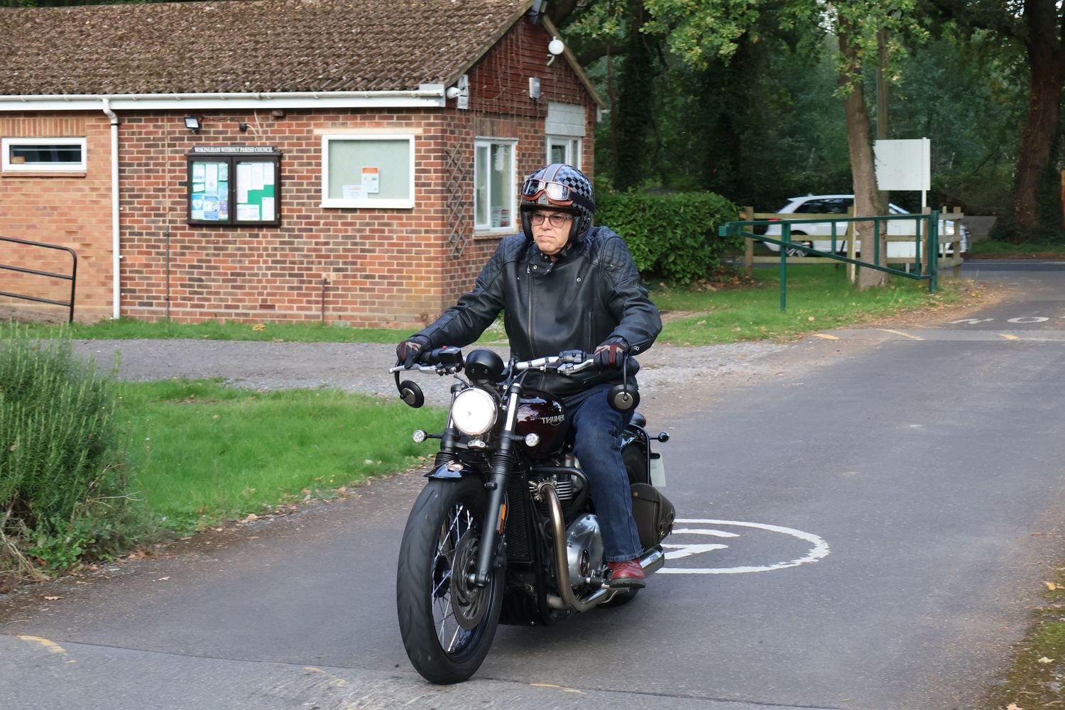 A motorcyclist rides a classic black motorcycle on a curved road past a brick building and green trees.
