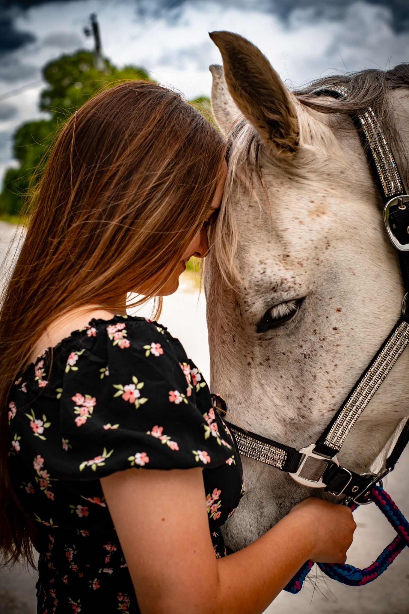 Person in floral dress shares tender moment with white horse against cloudy sky.