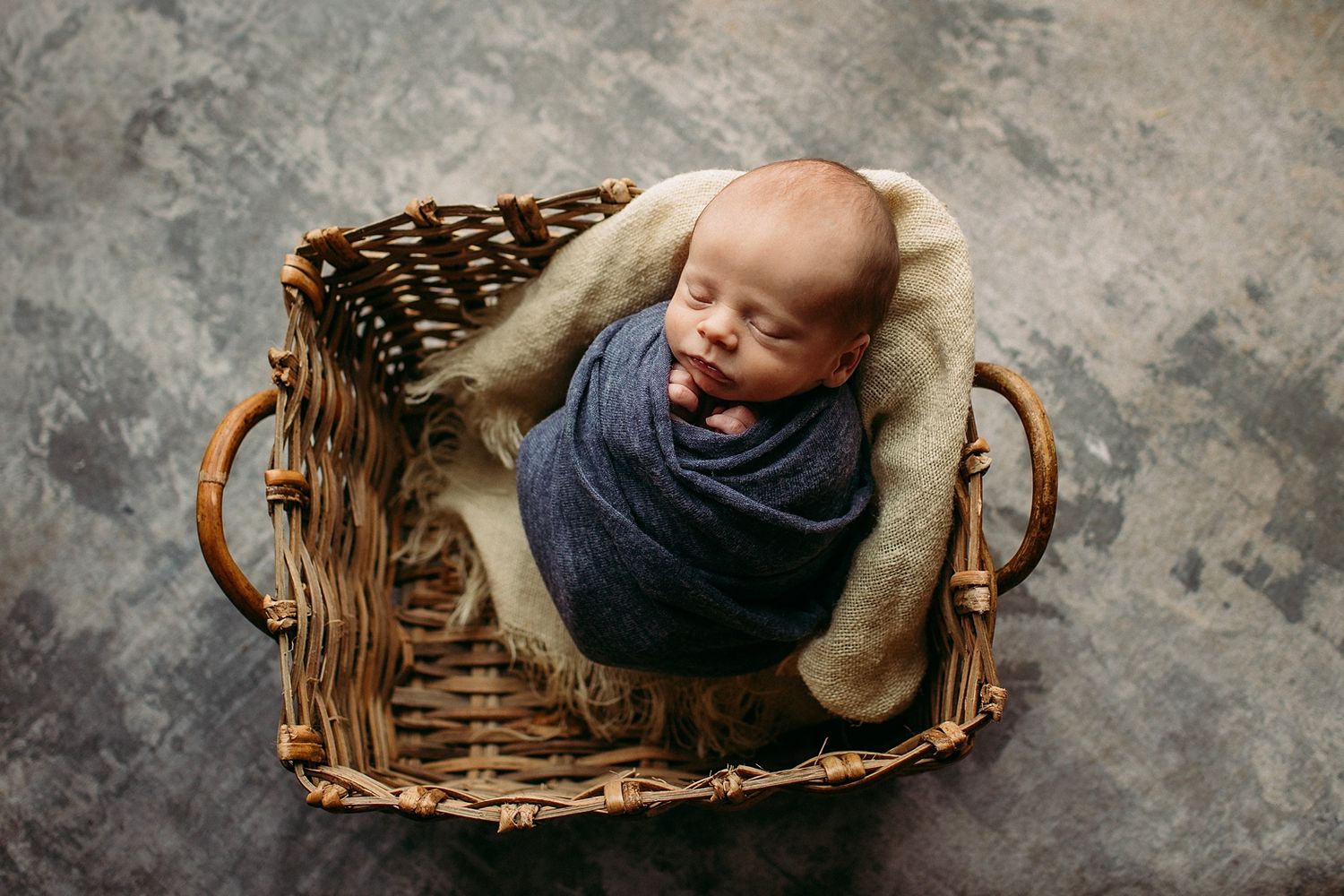A baby wearing a navy blue outfit lies curled up sleeping in a rustic woven basket on a textured gray background.