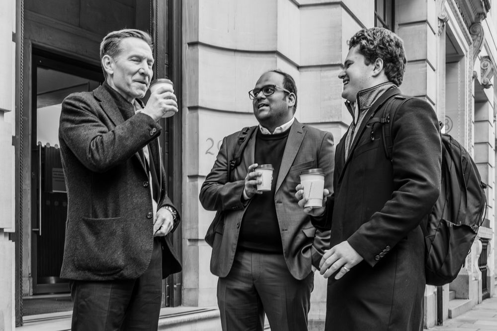 Three colleagues having a casual conversation while holding coffee cups outside an office building in black and white.
