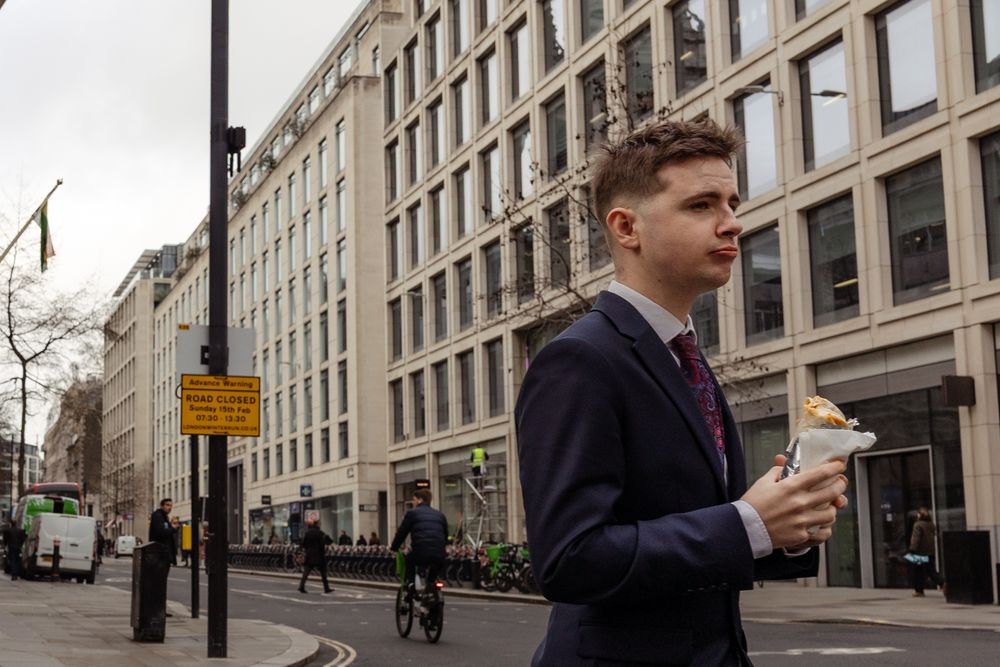 A businessman in a navy suit walks down a city street holding a coffee cup while passing historic office buildings.