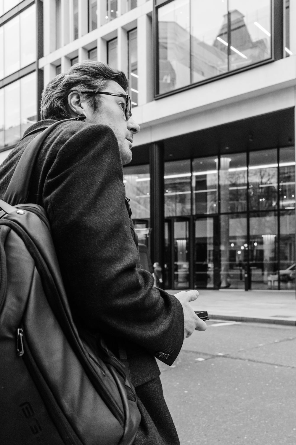 Black and white street photo of someone in glasses and winter coat carrying a backpack near modern buildings.