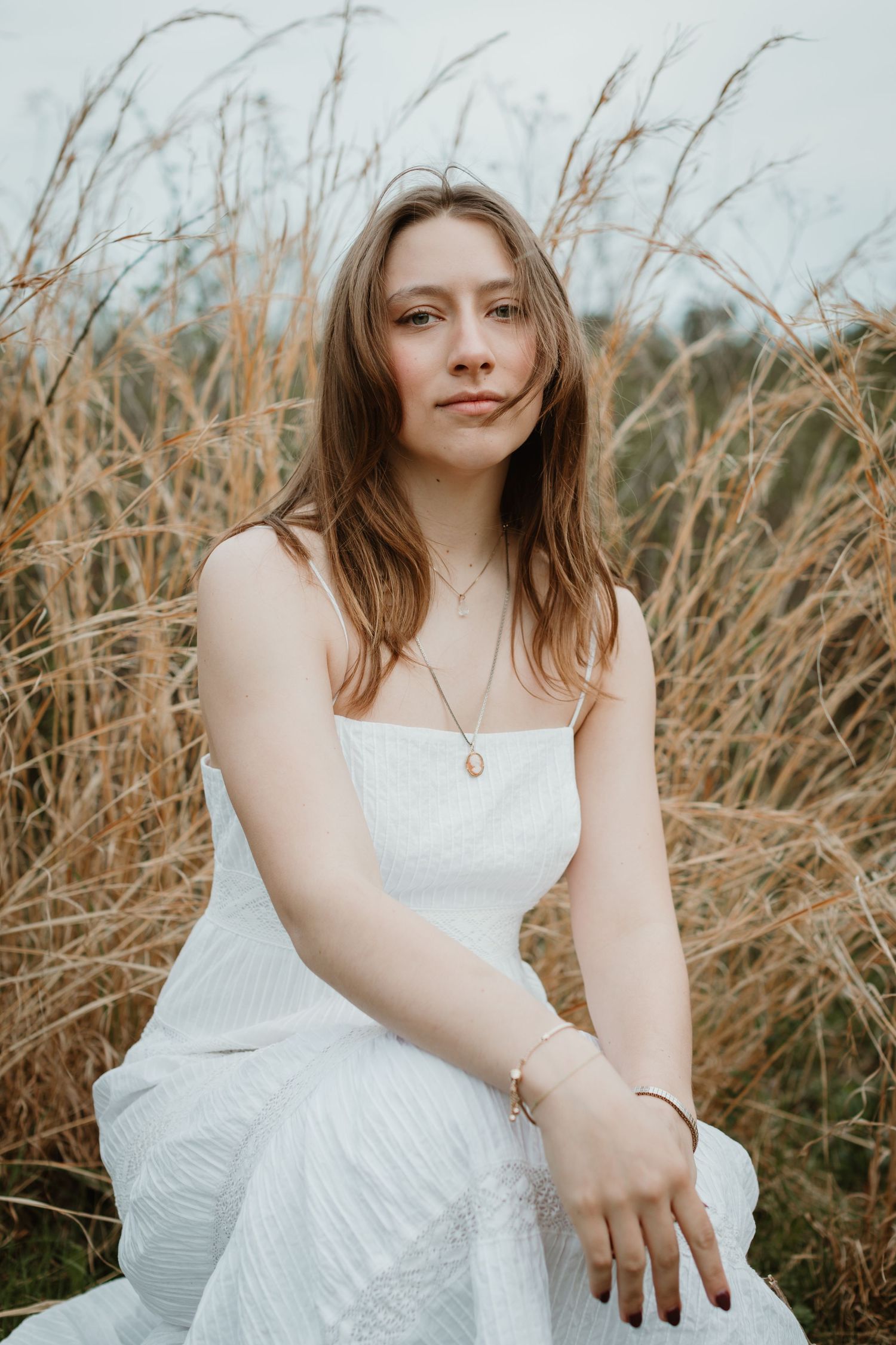 Young woman in white dress sitting gracefully among tall dried grasses at sunset.