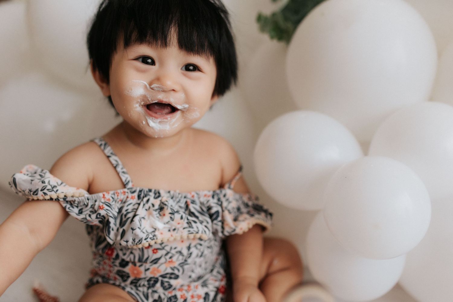 A baby in a floral dress smiles while sitting against a white backdrop with balloons.