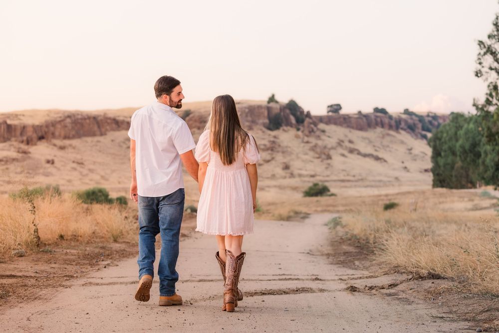 A couple walks hand in hand on a dirt path through a desert landscape during golden hour.