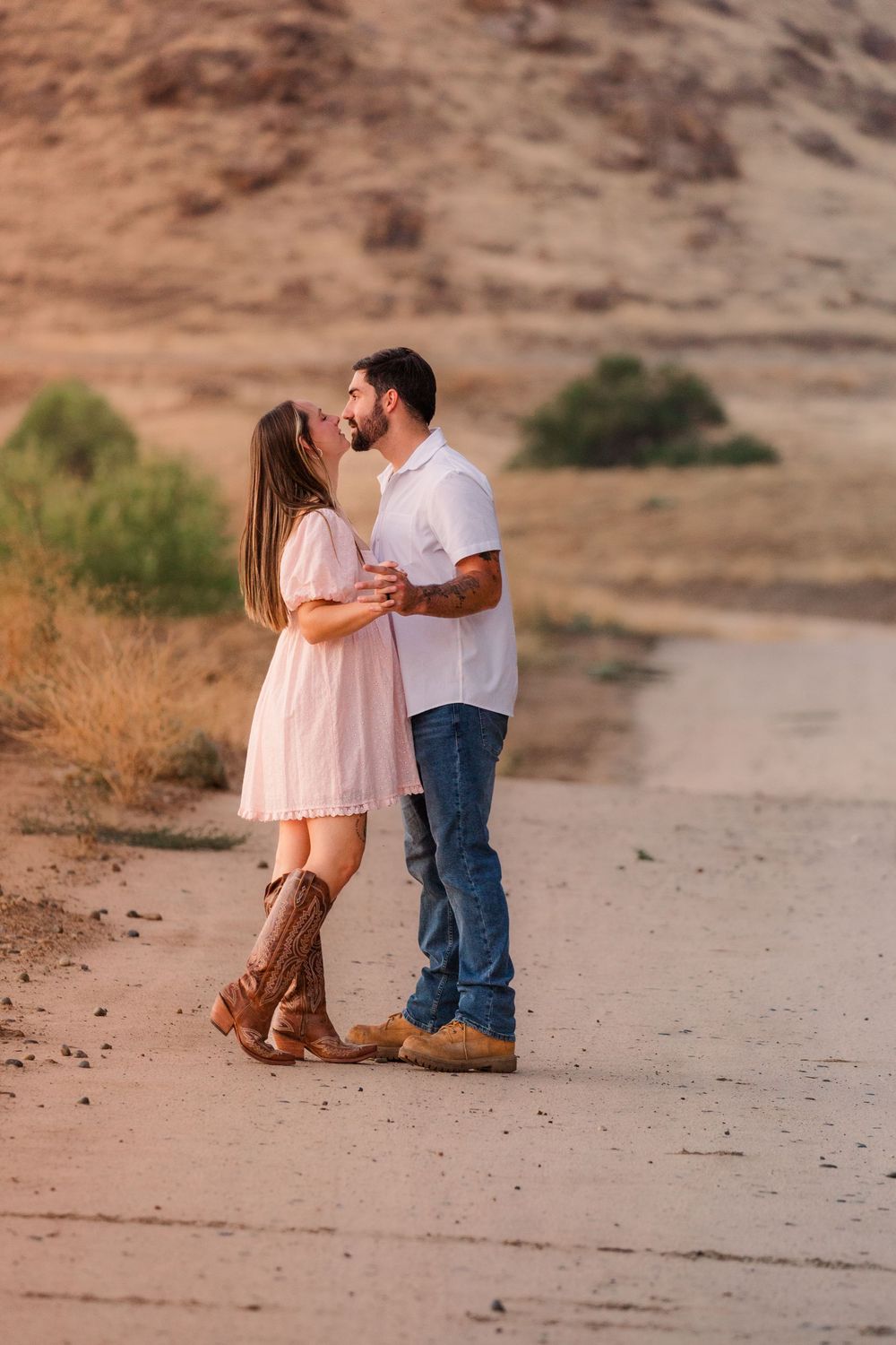 A couple shares a romantic moment in a desert landscape at sunset, wearing pink and white outfits.