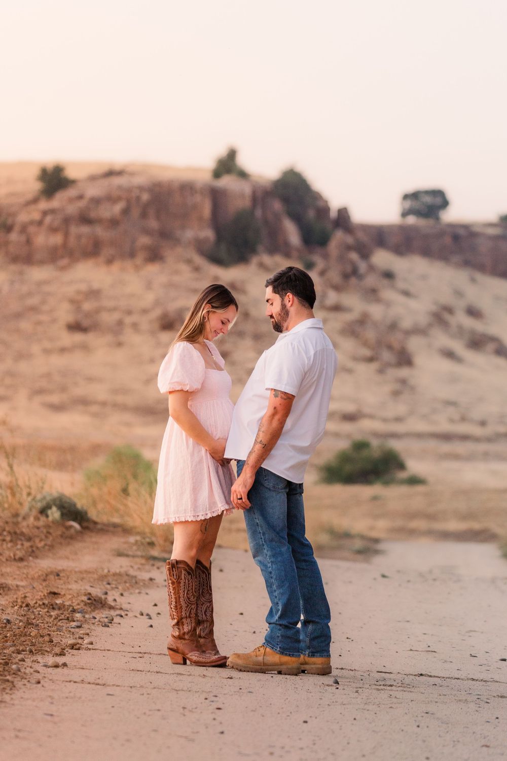 A couple stands together on a desert path during sunset wearing casual white tops and denim.