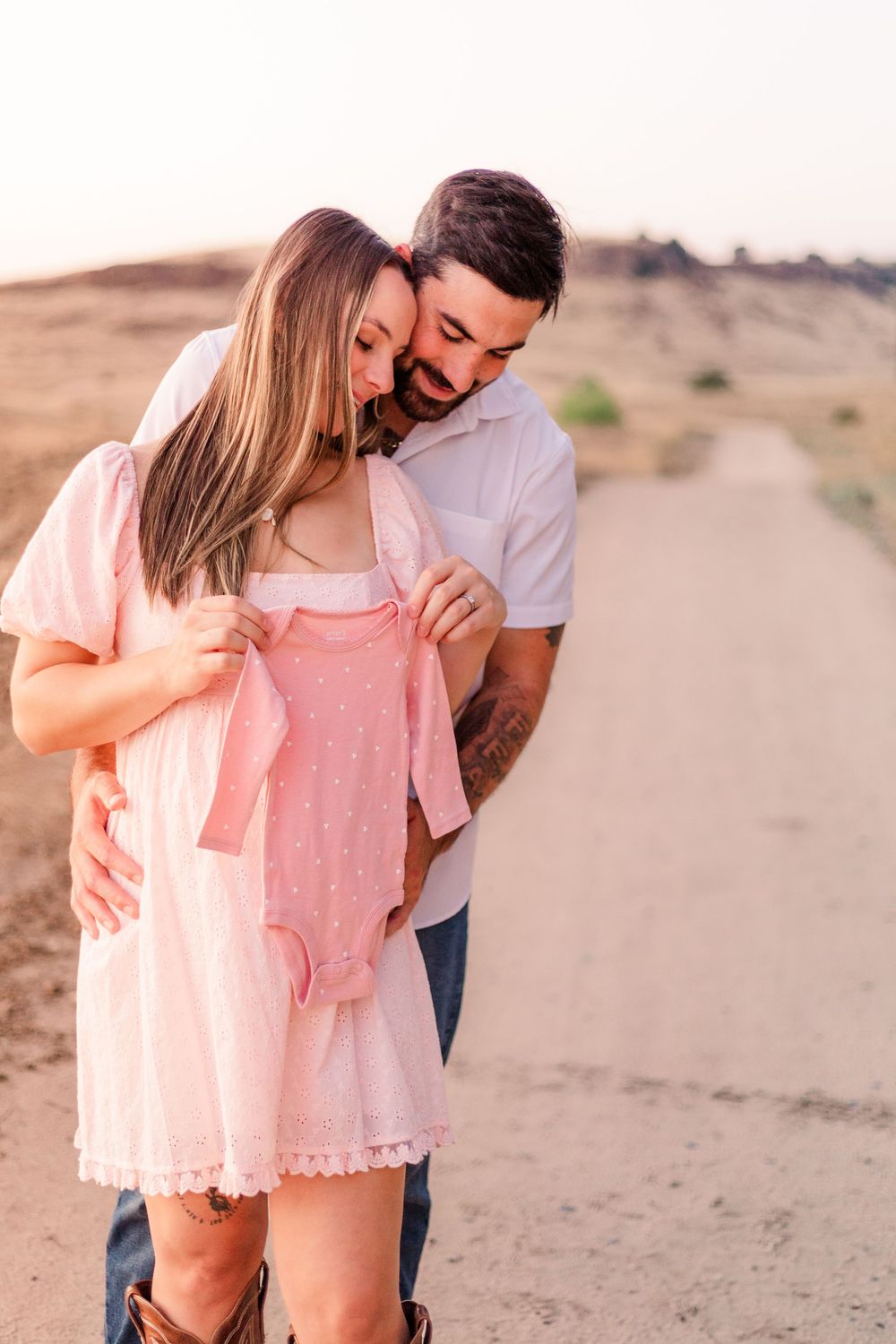 A couple embraces tenderly on a sandy desert path during a romantic photoshoot at sunset.