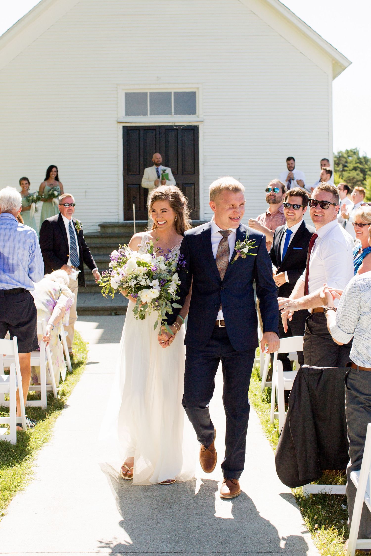 Wedding party exits white church building in summer sunshine while guests celebrate joyfully along walkway.