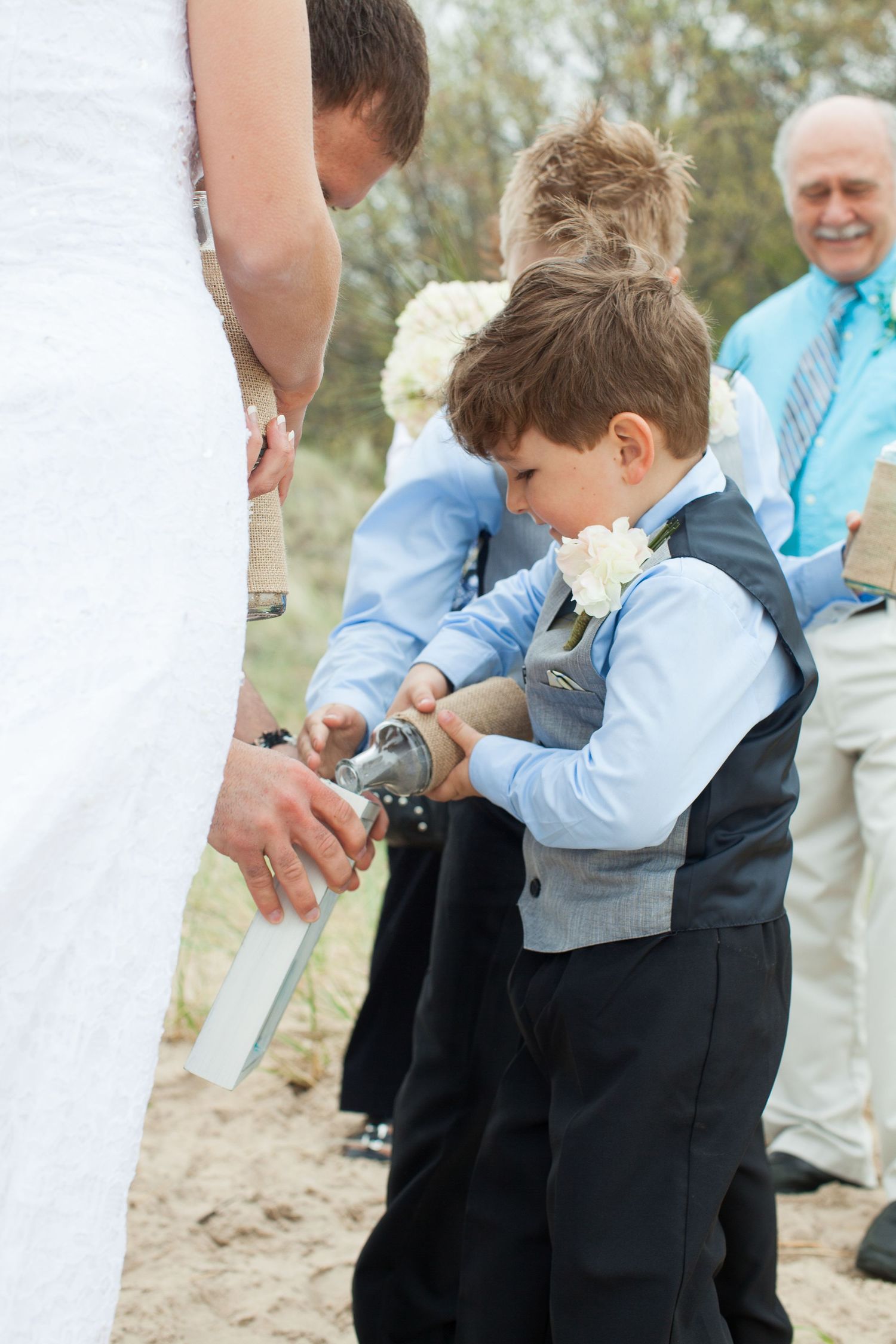 Young ring bearer in a blue dress shirt and black pants holds a pillow with rings during an outdoor wedding ceremony.