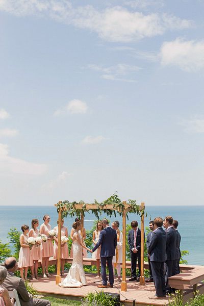 Outdoor wedding ceremony setup on a scenic oceanfront terrace with wooden arch and pink bridesmaid dresses.