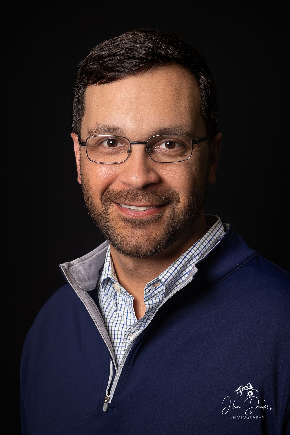 Professional headshot of person wearing navy quarter-zip and checkered shirt with glasses against dark backdrop.