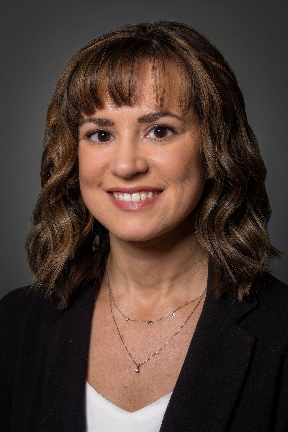 Professional headshot of person in black blazer with brown hair and warm smile against dark background.