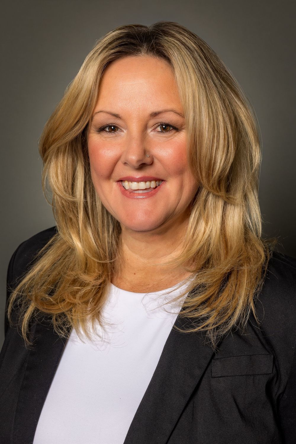 Business portrait of person wearing black jacket and white top with blonde hair against gray backdrop.