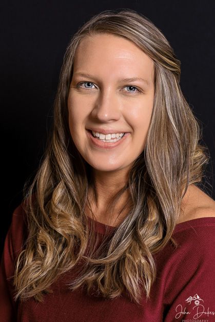 Studio portrait of person wearing burgundy top with wavy brown hair against dark background.
