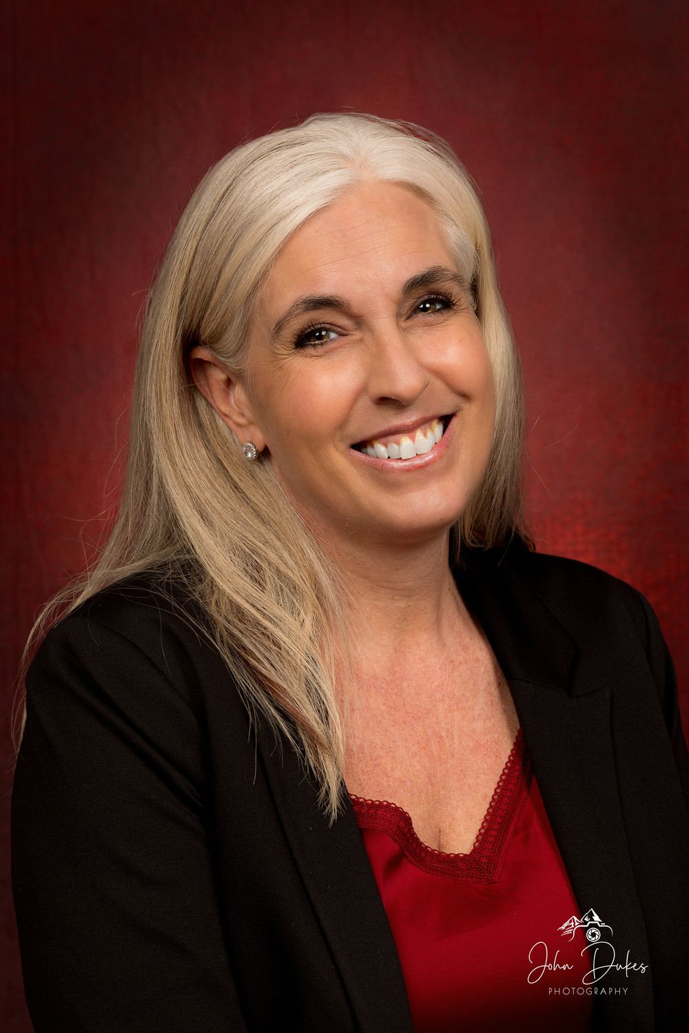 Professional headshot with person in black jacket and red top against burgundy backdrop.