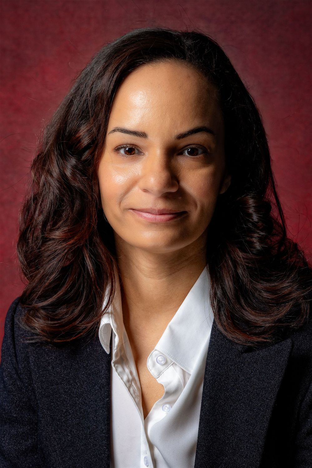 Professional headshot showing person in dark suit and white blouse against burgundy background.