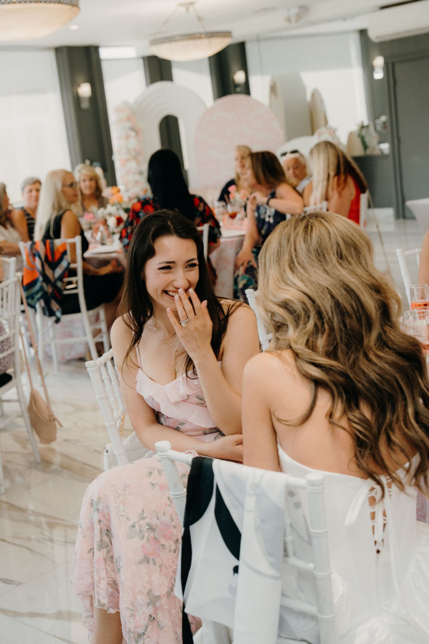 Guests in formal attire share a joyful moment at an elegant indoor event with white decor and natural lighting.