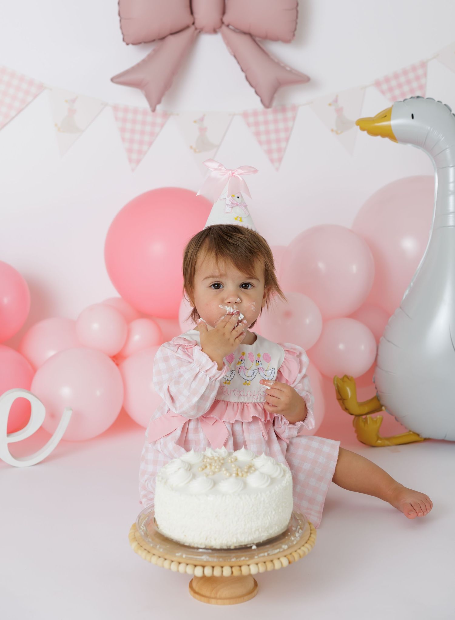 Pink balloon arch and bow backdrop with white cake on gold stand for birthday photos.
