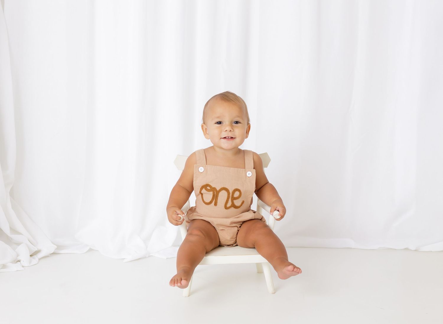 Baby in brown outfit sitting on white chair with 'one' text.