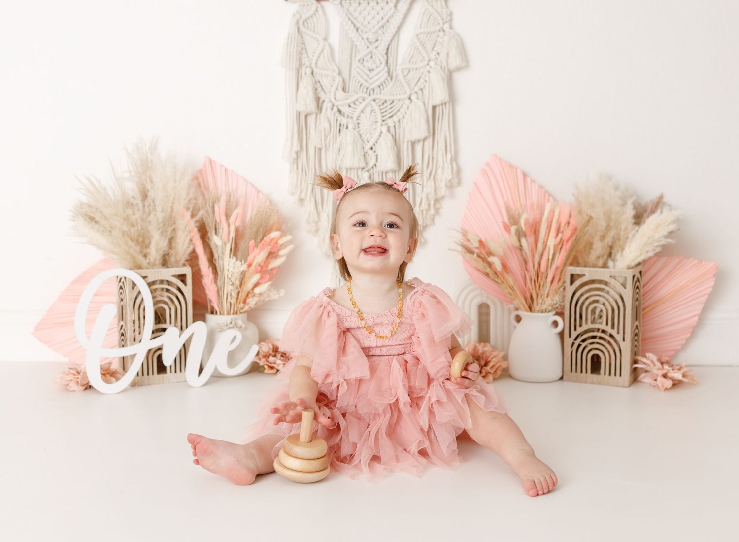 Baby in pink dress sitting with boho decorations and 'one' sign for first birthday.