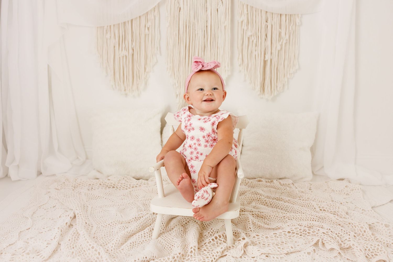 Baby in polka dot romper sitting on white chair against macrame backdrop.