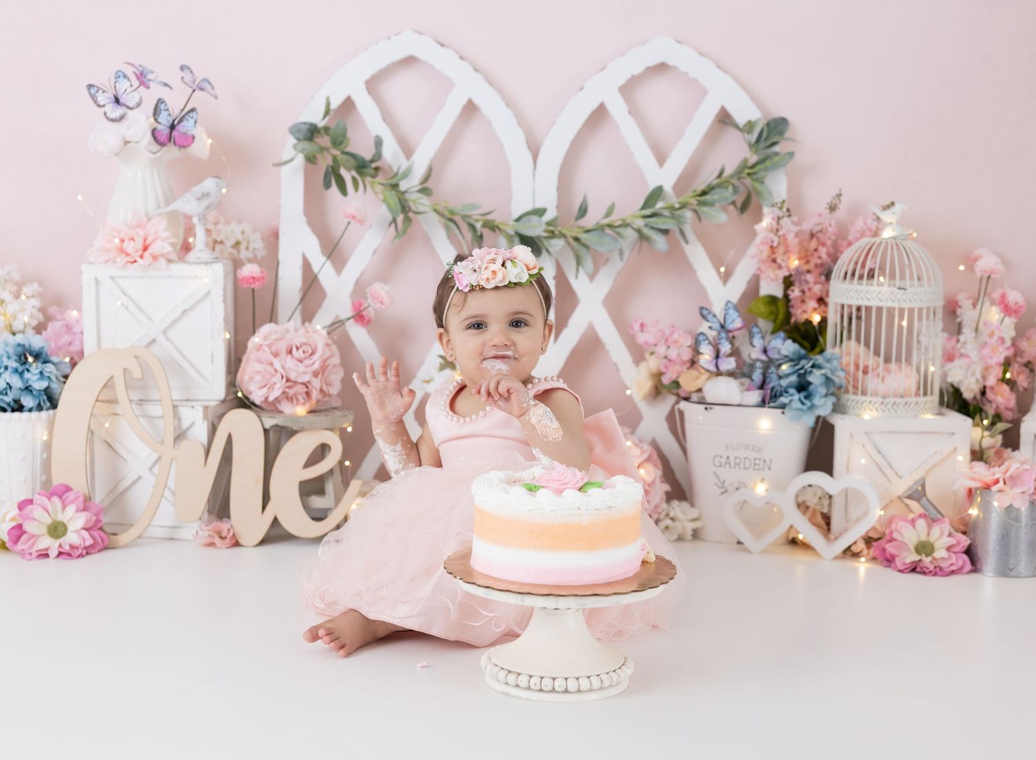 Romantic floral cake smash setup with pink backdrop, greenery and delicate decorations.