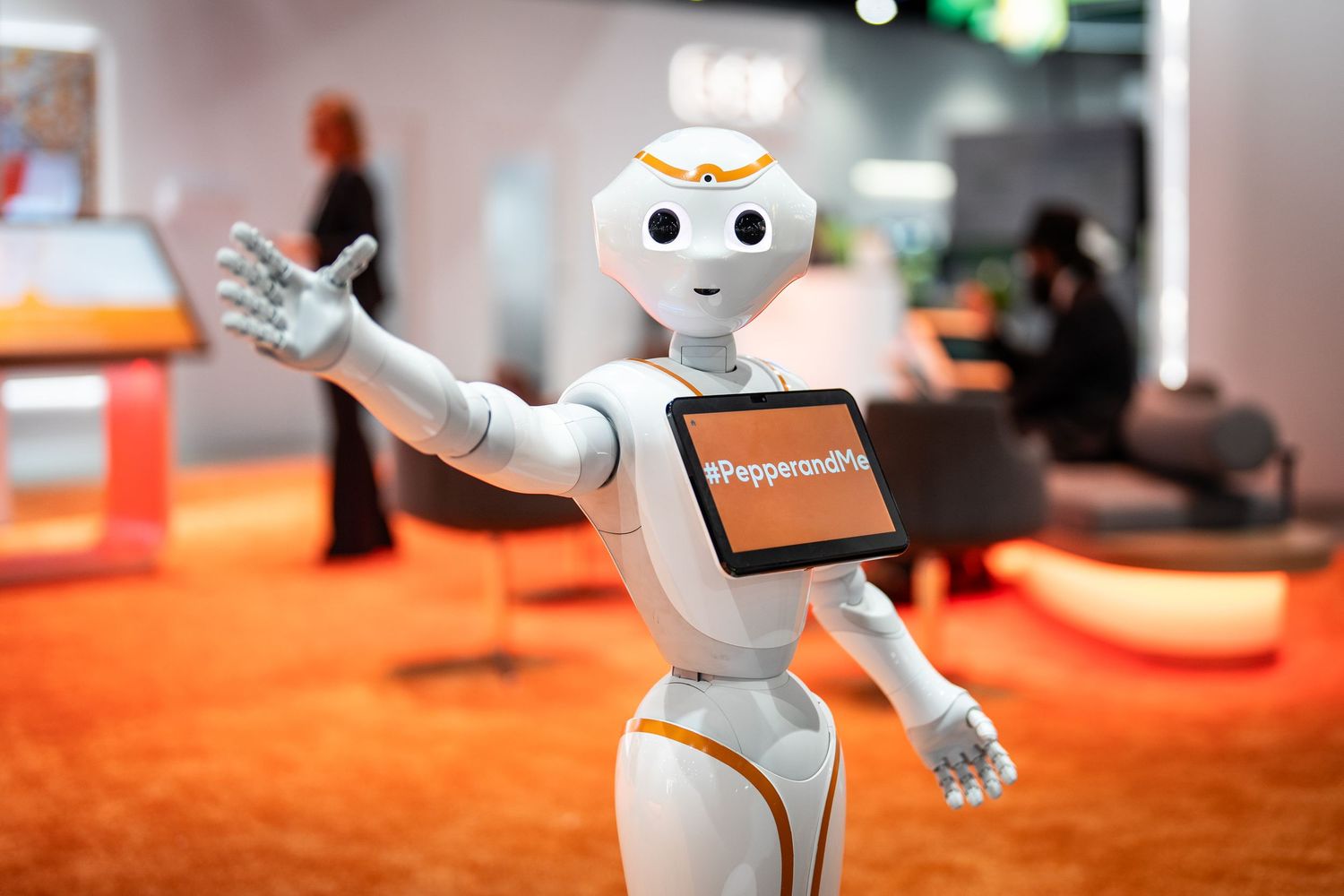 A white humanoid robot with expressive arms gestures on an orange carpeted floor at a technology exhibition.