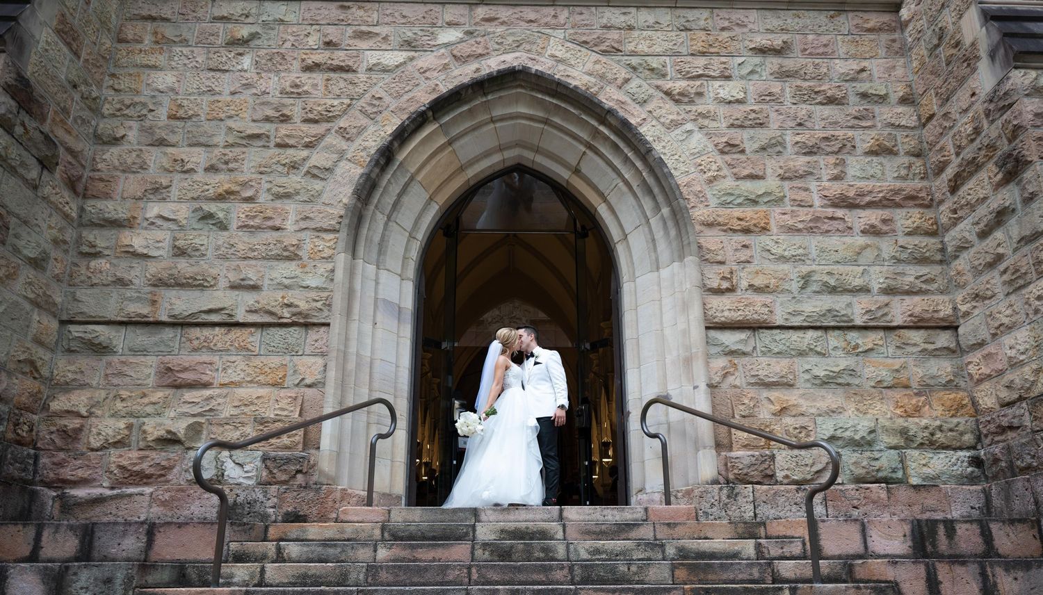 A newlywed couple shares a kiss on the steps of a stone church beneath a Gothic arched doorway.