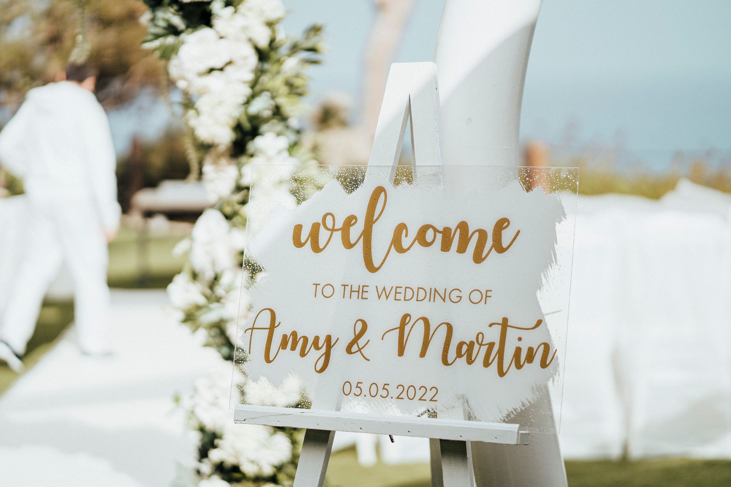 Elegant wedding welcome sign with gold calligraphy text displayed among white floral arrangements and greenery.