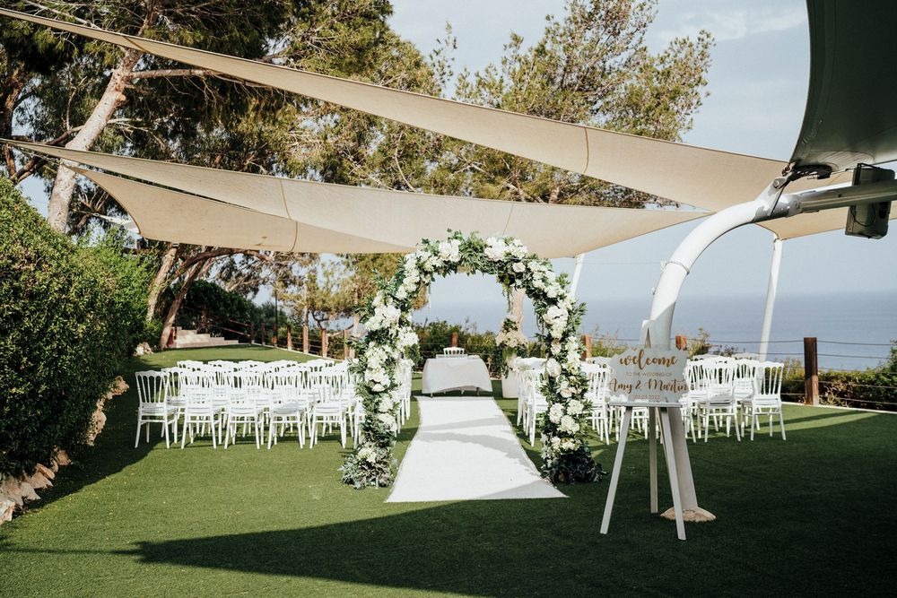 A panoramic view of an elegant outdoor wedding setup with white chairs and tents arranged on a lush green lawn.