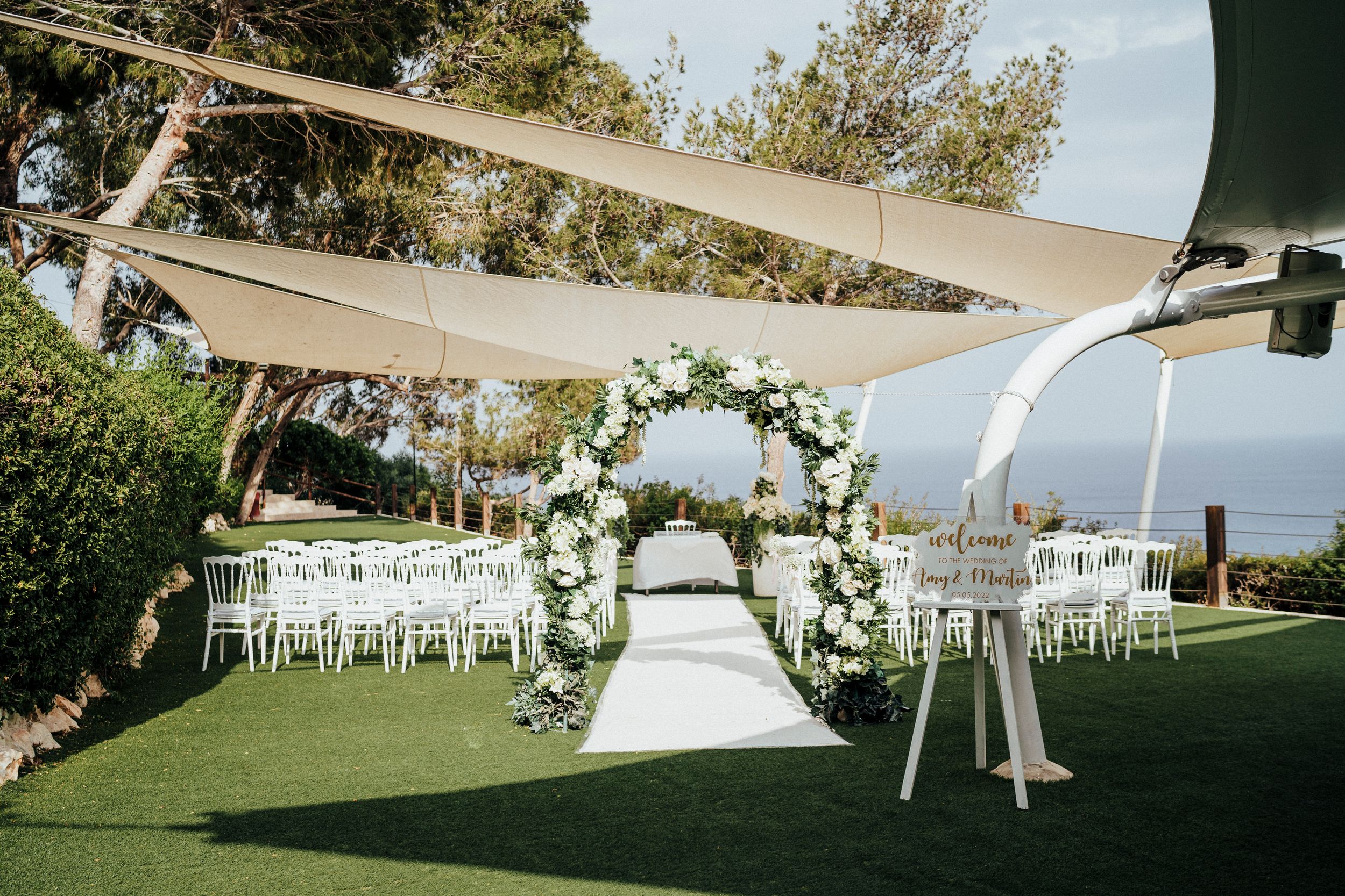 A panoramic view of an elegant outdoor wedding setup with white chairs and tents arranged on a lush green lawn.