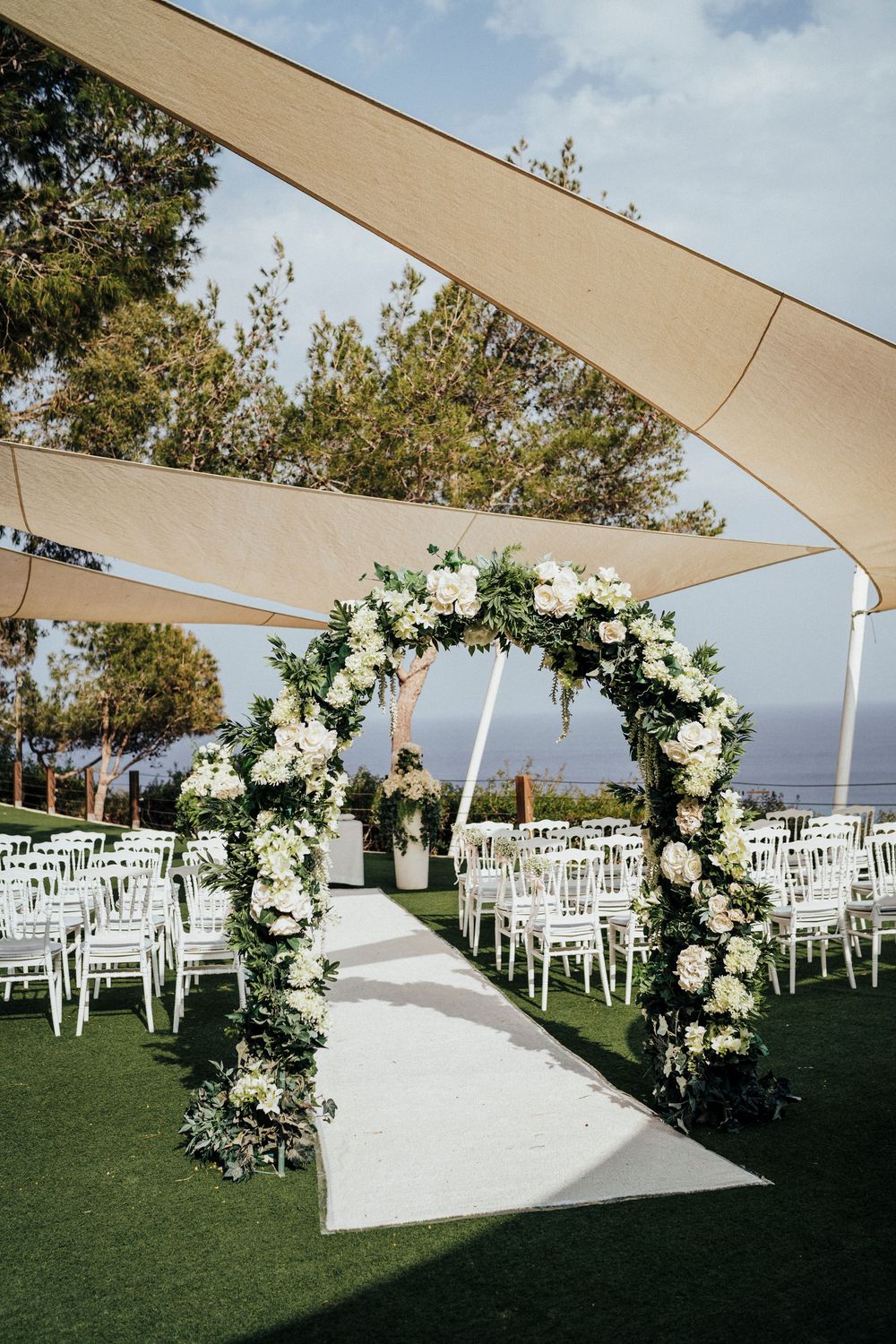 Elegant outdoor wedding ceremony setup with white floral arch, shade sails, and white chairs on manicured lawn overlooking ocean view.