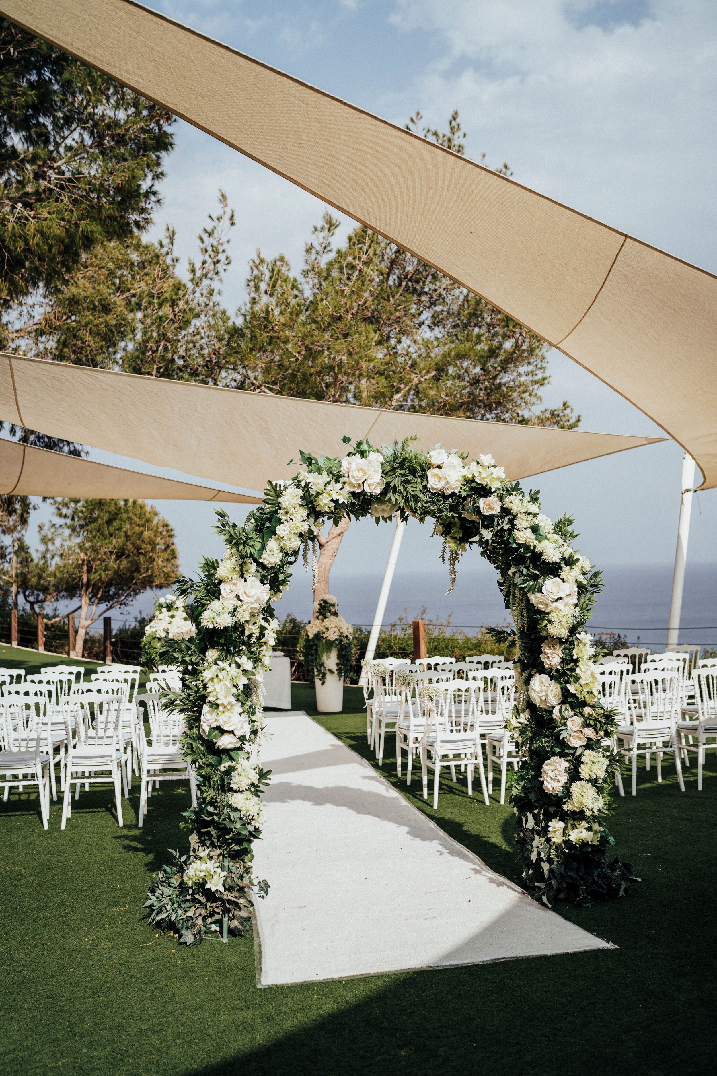 Elegant outdoor wedding ceremony setup with white floral arch, shade sails, and white chairs on manicured lawn overlooking ocean view.