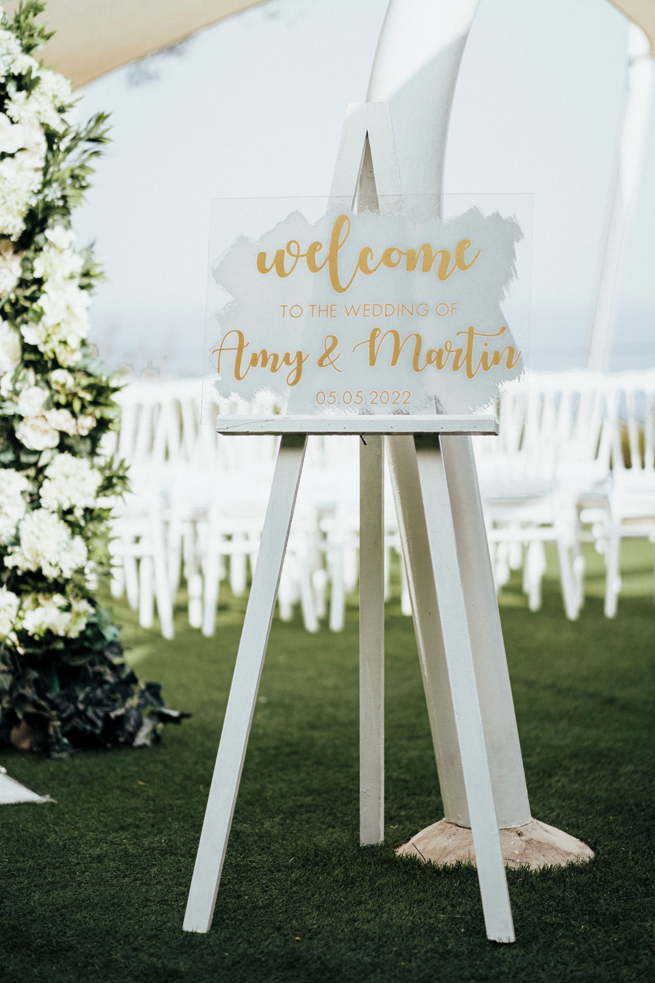 Elegant white wedding welcome sign on easel with gold lettering and floral decorations in an outdoor garden setting.