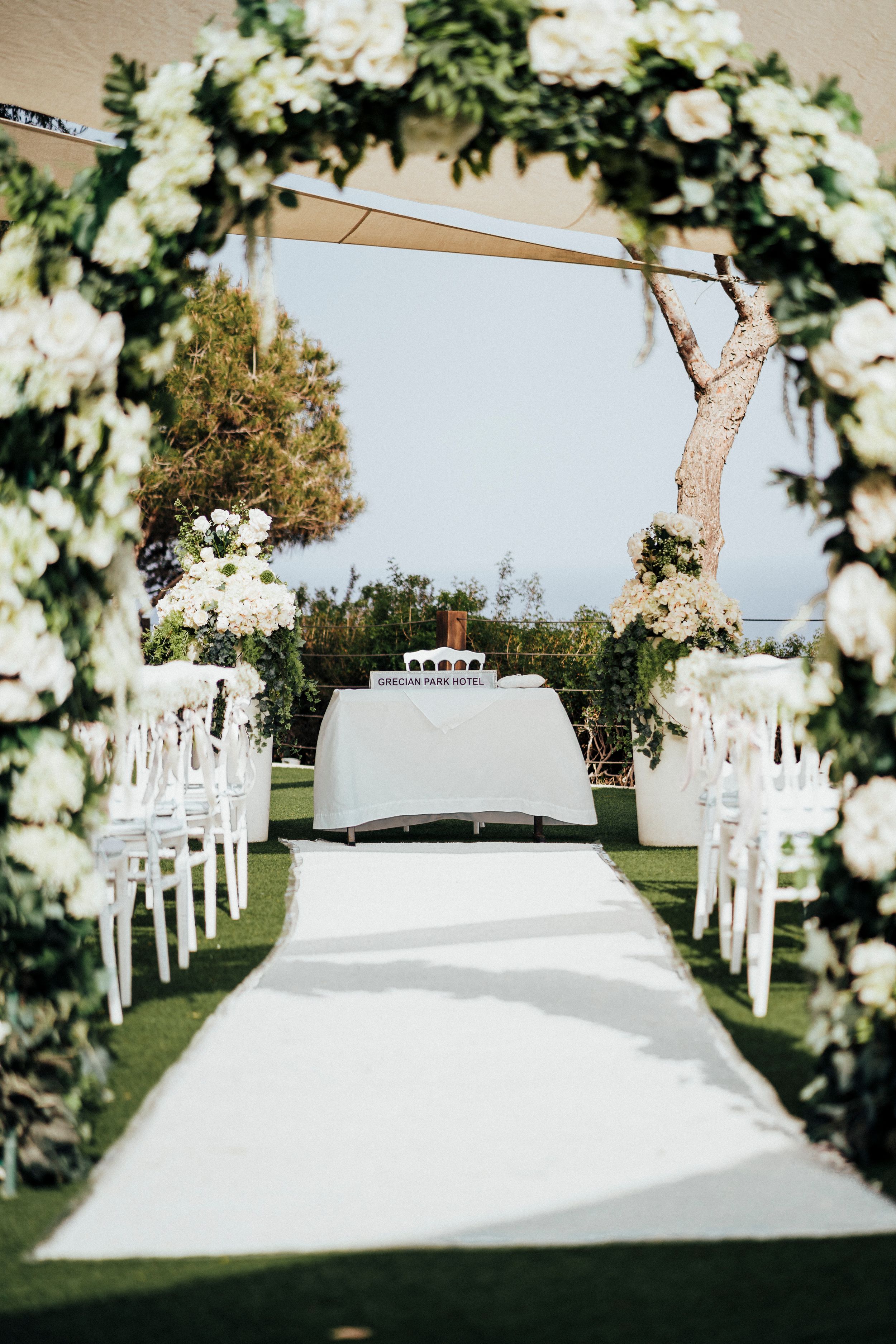 White chairs arranged in rows for an outdoor wedding ceremony with floral arch and white aisle runner.