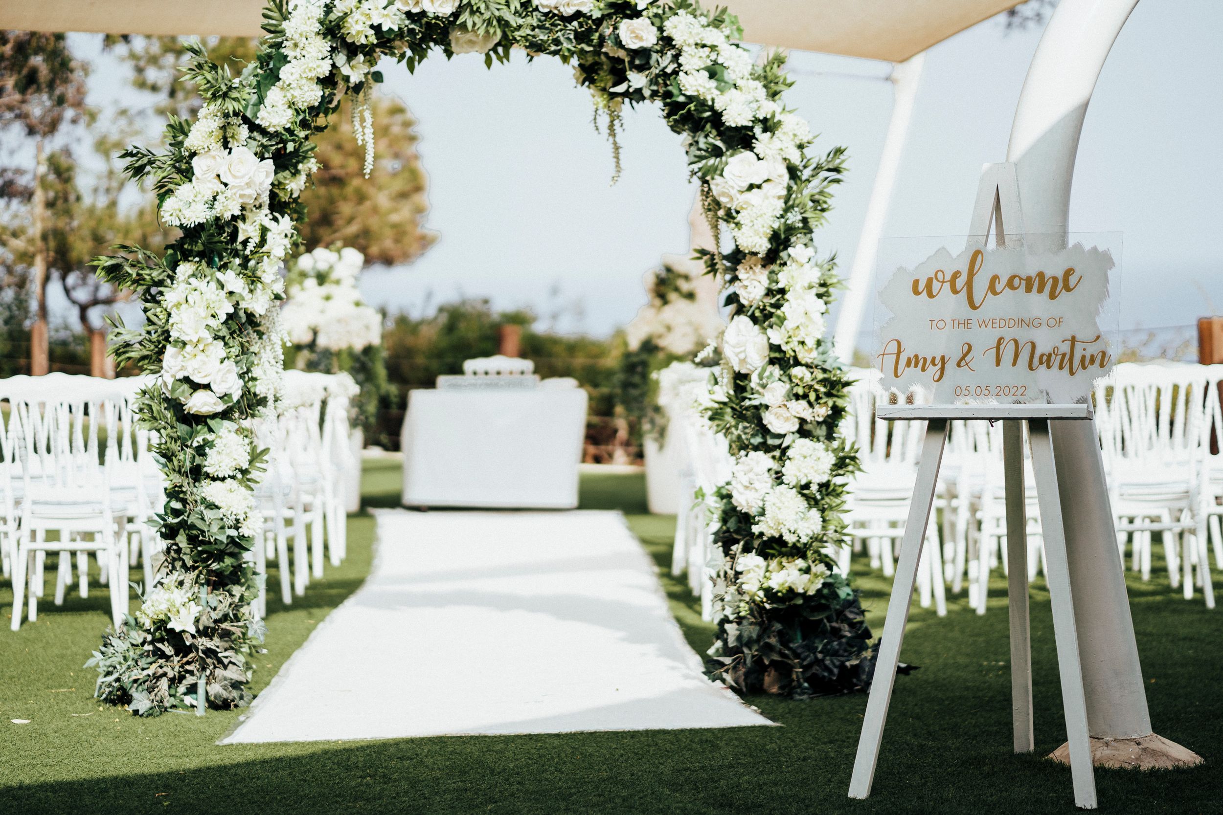 Elegant outdoor wedding ceremony setup with white floral arch and chairs decorated with greenery and white flowers.
