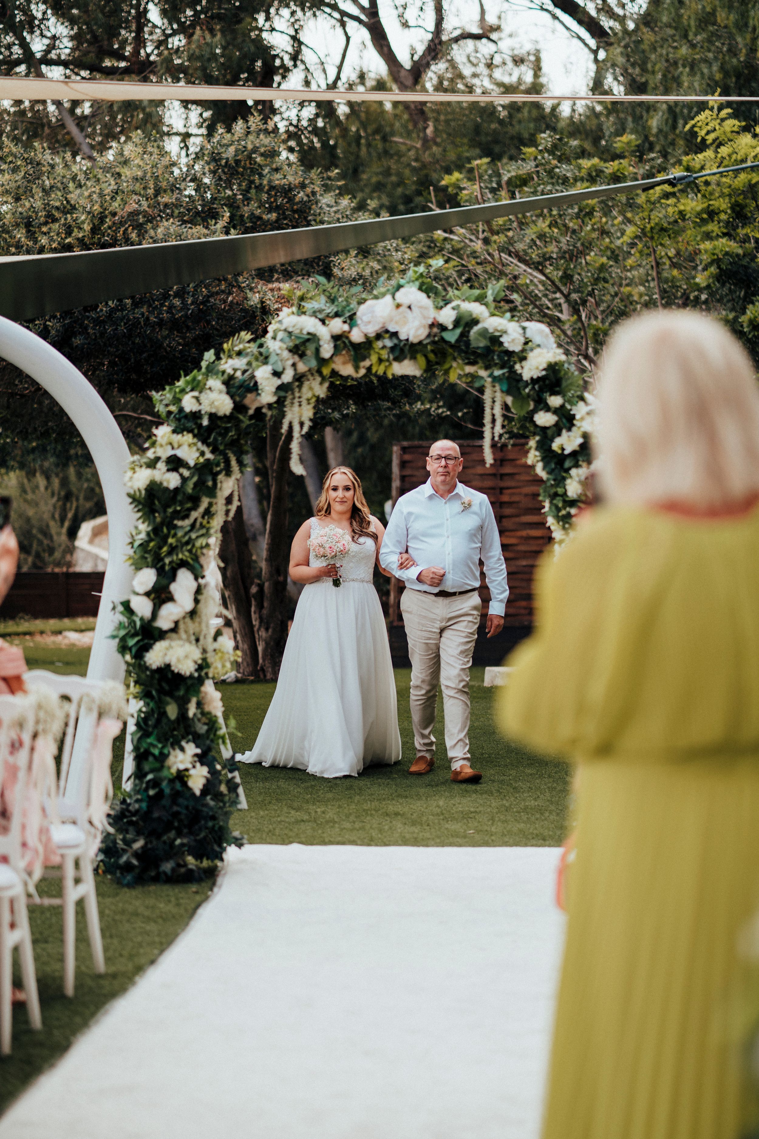 Outdoor garden wedding ceremony setup with white floral arch and guests seated on white chairs in natural setting.