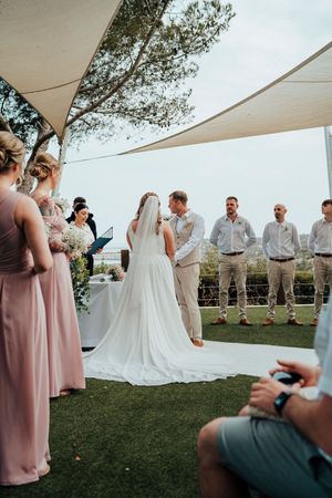 Outdoor wedding ceremony under white canopy tent with bridesmaids in pink dresses and groomsmen in light attire.