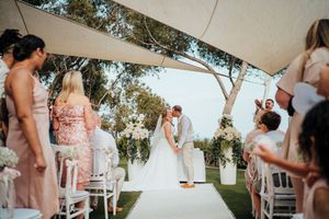 Outdoor wedding ceremony setup with white chairs and floral arrangements under natural sunlight.