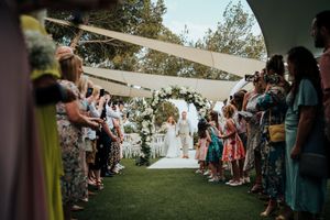 Wedding guests line both sides of a walkway under a white tent canopy during an outdoor ceremony.