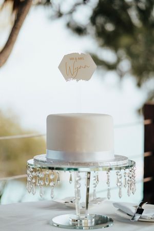 White wedding cake displayed on an elegant crystal stand with hanging crystal decorations against a light background.
