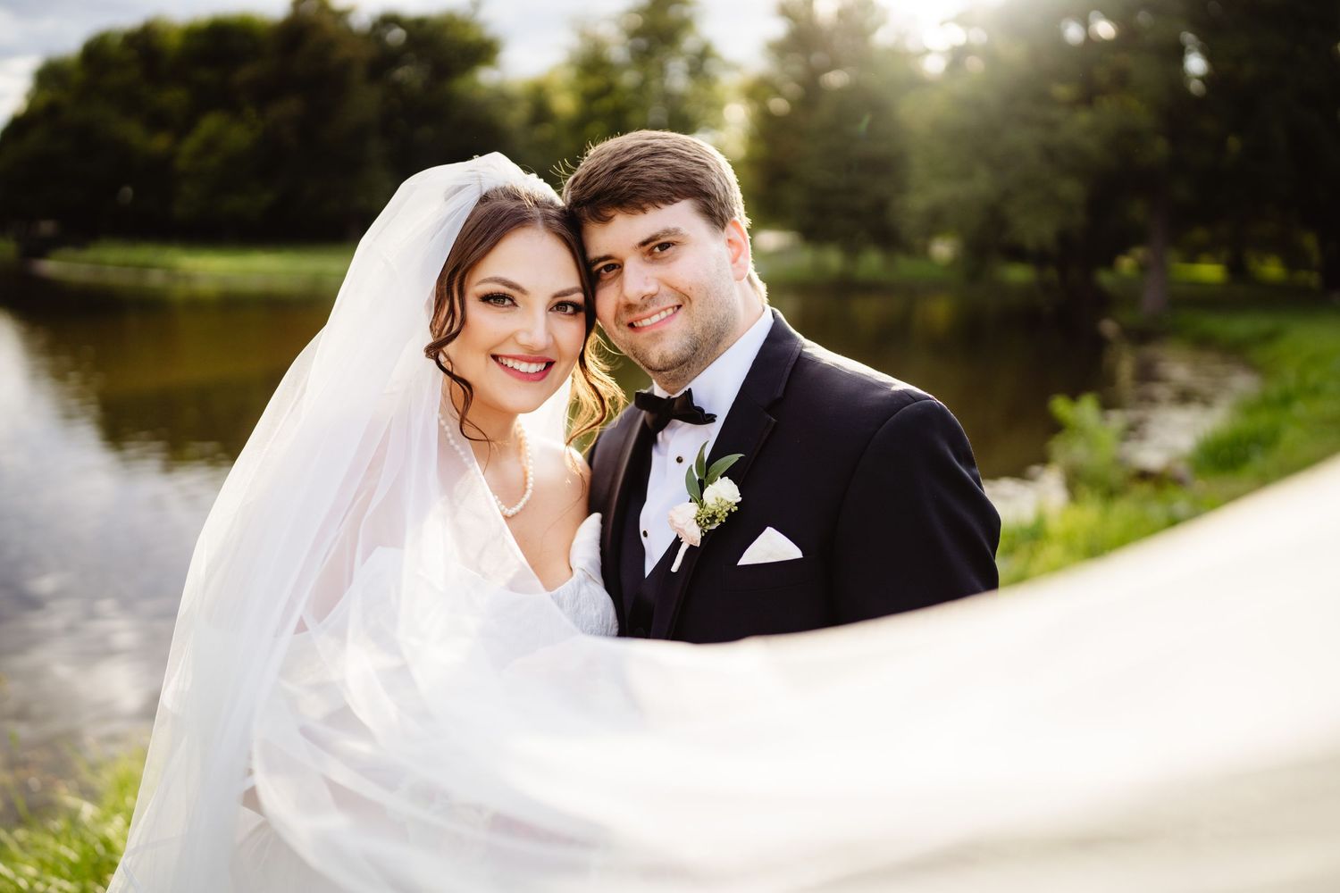 Elegant bride and groom posing together outdoors near a lake on their sunny wedding day.