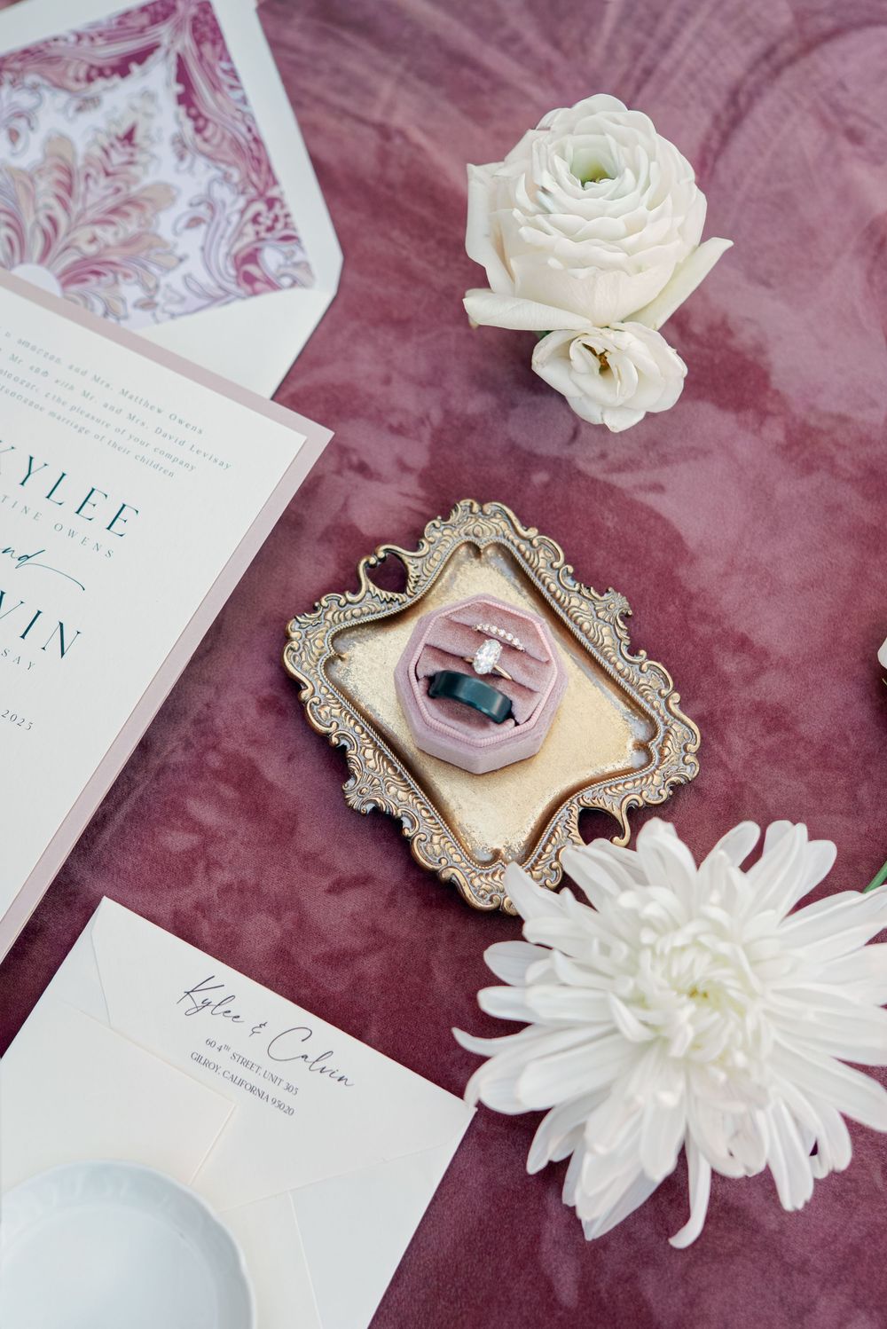 Wedding details flatlay with white flowers, invitation and ornate frame on burgundy velvet.
