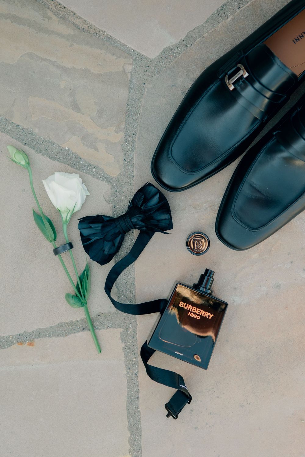 Wedding accessories including blue dress shoes, bow tie, cologne bottle and white flower on stone floor.