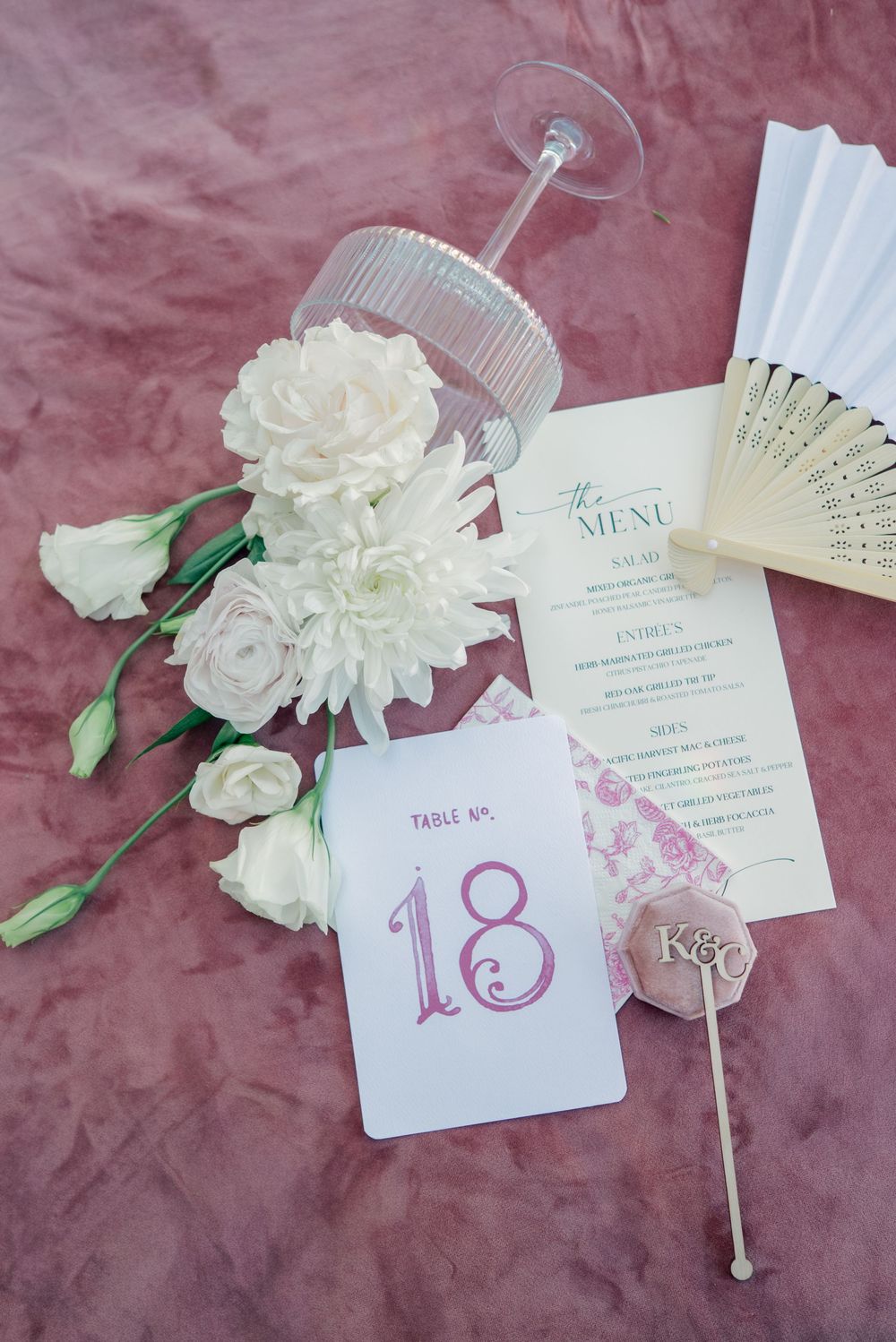 Wedding stationery including menu cards, table numbers, and white floral decorations arranged on pink velvet fabric.