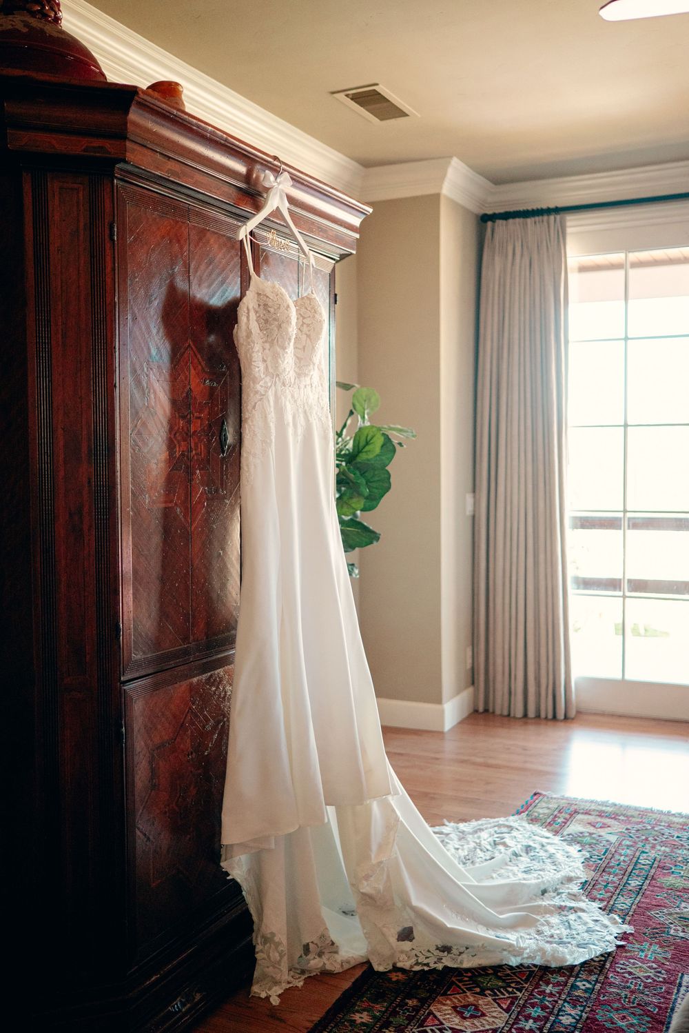 A white wedding dress hangs elegantly against a dark wooden armoire in a bright room with natural lighting.
