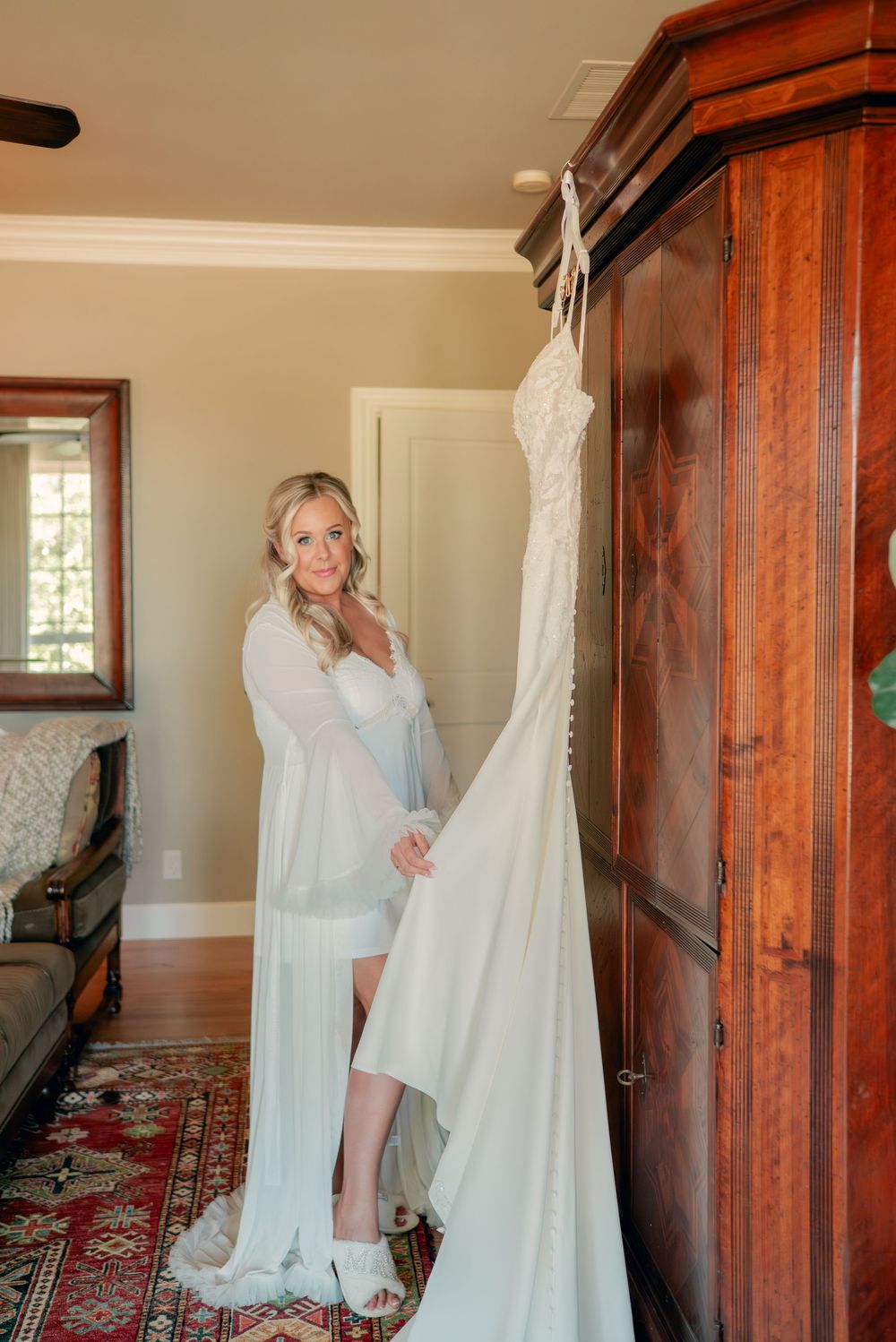 White wedding dress displayed on a wooden wardrobe while being admired in a sunlit room.