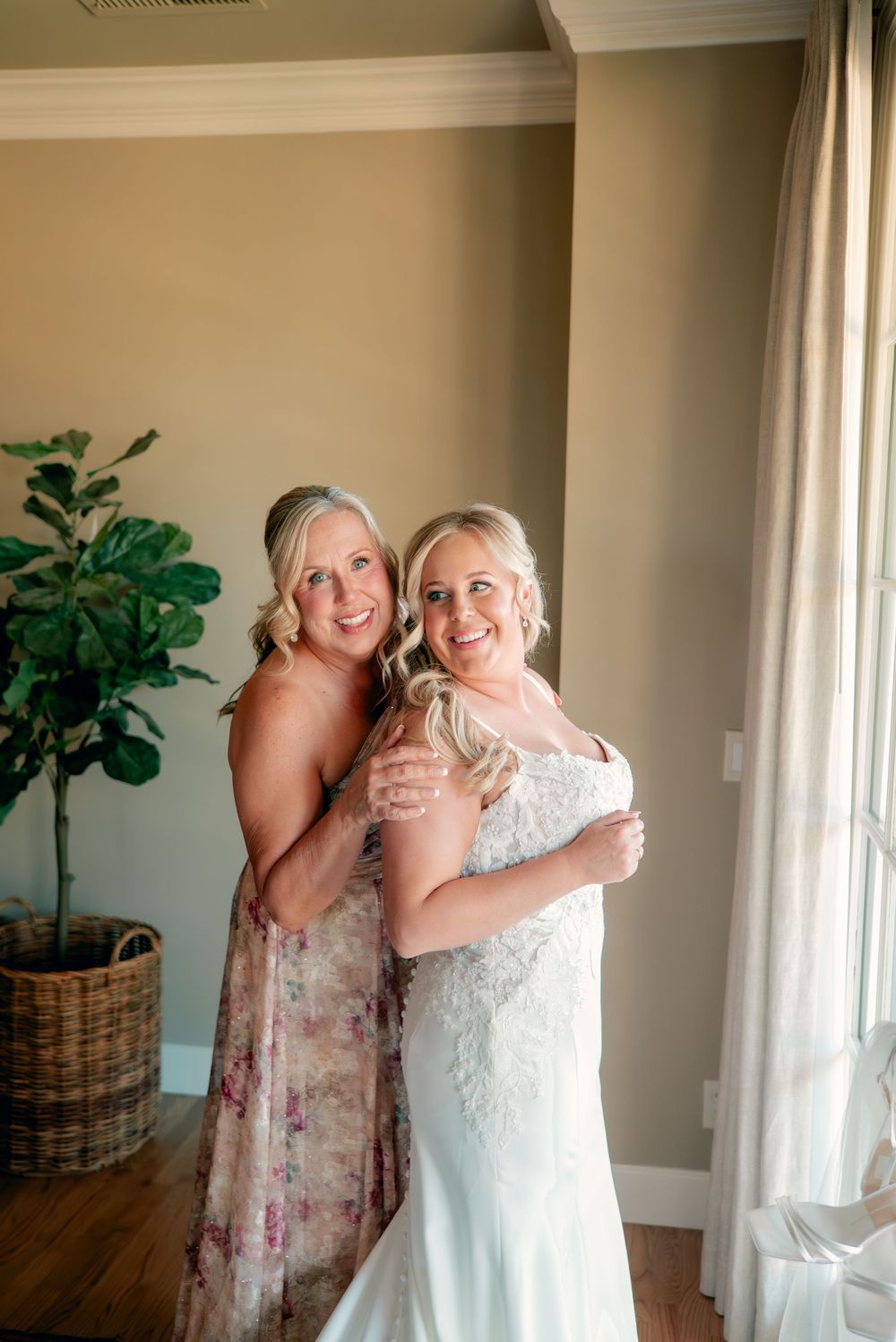 Two women embrace in a sunlit room with a potted fiddle leaf fig plant and white curtains.