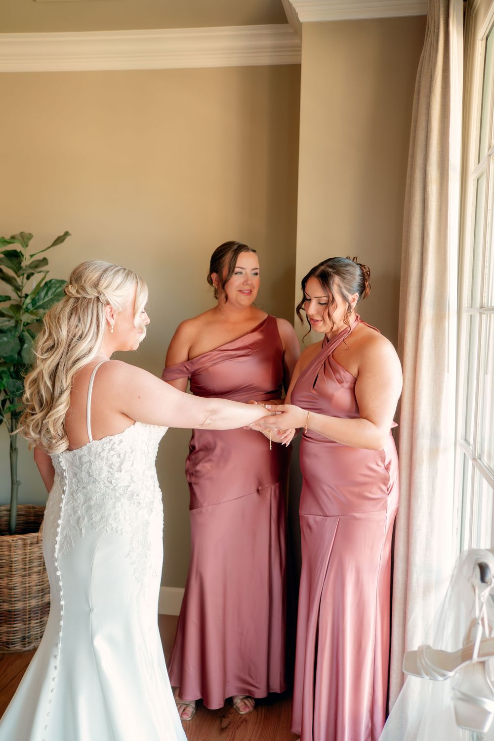 Wedding party members in pink bridesmaid dresses help with final preparations in a sunlit room.